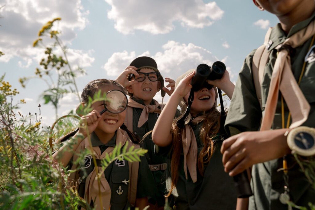 Group of children exploring nature at a summer camp, wearing scouting uniforms and using binoculars.